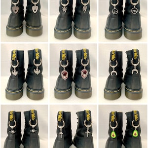 May include: Nine pairs of black leather boots with silver charms attached to the laces. Charms include hearts, a fairy, an anchor, a gemstone, a crescent moon, a cross, and an avocado. The boots have yellow stitching and the text "Air Wair" on the back.