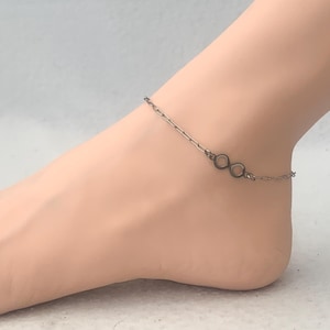 May include: A silver-coloured anklet with an infinity symbol charm. The fine chain is draped around the ankle, highlighting the minimalist design. The anklet is a simple yet elegant piece of jewellery.