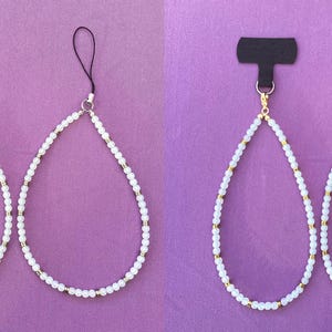 May include: Two beaded phone charms with a teardrop design. Each charm features white beads and gold-colored accents, connected to a black tab and a black cord. The charms are displayed against a purple background.