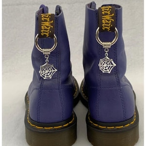 May include: Purple leather Dr. Martens boots with black laces and yellow stitching. The boots have a spiderweb charm attached to the laces. The boots have a black and yellow "Dr. Martens" tag on the back.