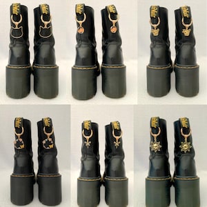 May include: Six pairs of black leather boots with gold charms attached to the laces. The charms feature various designs, including a cat, a sun, and a dragonfly.