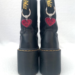 May include: Black leather platform boots with yellow stitching and a thick platform sole. The boots have a silver charm attached to each boot that says "Bite Me" on a red heart.