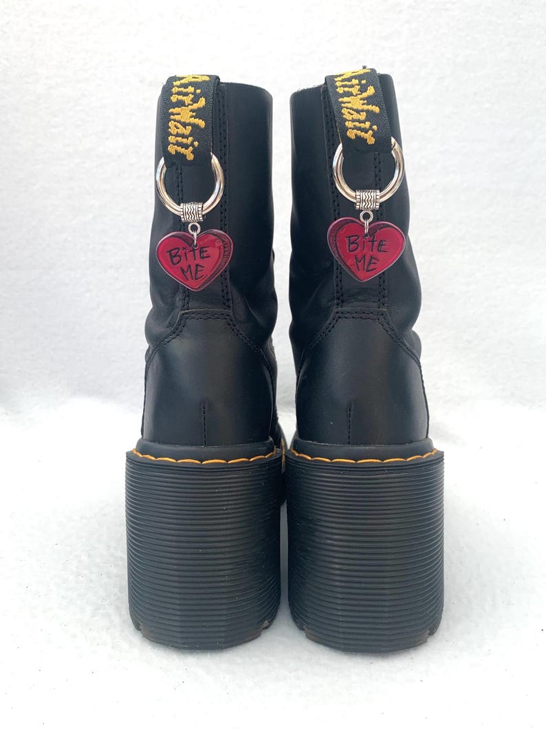 May include: Black leather platform boots with yellow stitching and a thick platform sole. The boots have a silver charm attached to each boot that says "Bite Me" on a red heart.