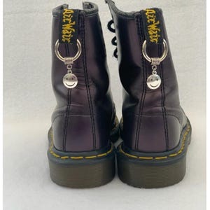 May include: A pair of black leather Doc Martens boots with yellow stitching and a smiley face charm attached to each boot. The boots have a black lace-up closure and a black rubber sole.