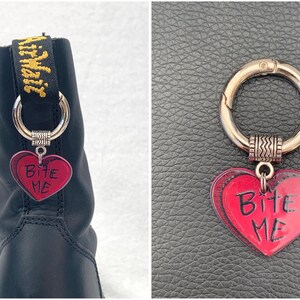 May include: A black boot with a silver charm attached to the laces. The charm is a red heart with the words "Bite Me" written on it.