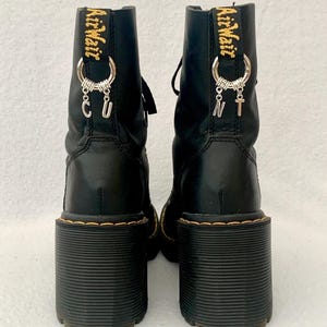 May include: Black leather platform boots with chunky heels and gold AirWair tags. The boots have silver charms attached to the tags, spelling out "CUNT".