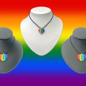 May include: Three necklaces with rainbow heart pendants on black cords. The pendants are displayed on grey and white bust forms against a rainbow gradient background. The heart pendants feature the colours of the rainbow.