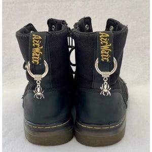 May include: Black canvas high-top boots with black laces and a zipper on the side. The boots have a spider charm attached to a silver ring on each boot. The boots have the text "Azz MAgic" embroidered on the back of each boot.