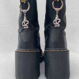 May include: Black leather boots with a chunky heel and a snake charm attached to the laces. The boots have a yellow and black "AirWair" tag on the back.