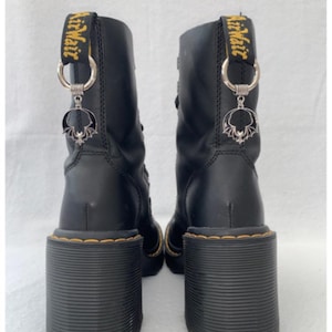 May include: Black leather boots with a chunky heel and a yellow "AirWair" tag. The boots have a silver charm with a bat design attached to the tag.