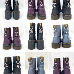 May include: Nine pairs of black leather boots with different coloured accents and charms. The boots have a lace-up front and a yellow stitching detail. The charms are attached to the laces and include a variety of designs, such as a spider, a crescent moon, a cross, and a fairy.