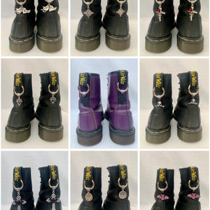 May include: Nine pairs of black and purple leather boots with yellow stitching and Air Wair text. Each boot has a silver charm attached to the back loop. Charms include crosses, bats, and other decorative elements.