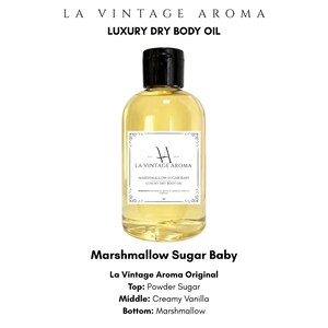 May include: A clear plastic bottle filled with golden-yellow liquid body oil, featuring a black flip-top cap. The white rectangular label displays the text LA VINTAGE AROMA, MARSHMALLOW SUGAR BABY, and LUXURY DRY BODY OIL with decorative border accents.