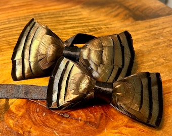 May include: Two feather bow ties. The bow ties are made of brown and gold feathers with black satin centers and brown straps. The bow ties are displayed on a wooden surface.
