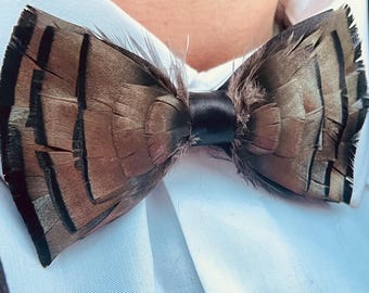 May include: A brown feather bow tie with black accents, tied with a black satin ribbon. The bow tie is set against a white dress shirt and a dark jacket, creating a formal look.
