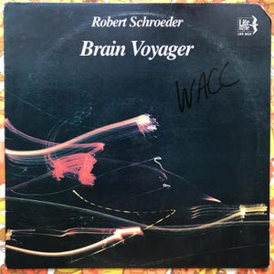 May include: A black vinyl record album cover with the title "Brain Voyager" by Robert Schroeder. The album cover features abstract lines of light in yellow, red, and white against a black background. The album cover is signed "WACC" in black marker.
