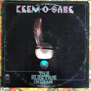 May include: A black vinyl record album cover with the title "Kemo-Sabe The Electric Indian" in colorful text. The cover art features a stylized image of a Native American headdress with a feather.