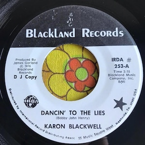 May include: A black and white vinyl record with a yellow and green flower design. The record label is white with black text that reads "Blackland Records", "IRDA # 253-A", "Time 3:15", "Blackland Music Company, Inc. BMI", "DANCIN' TO THE LIES (Bobby John Henry)", "KARON BLACKWELL", and "Distributed by: International Record Distributing Assoc 55 Music Square West Nashville, Tn. 37203 USA".