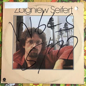 May include: A vinyl record album cover featuring a photo of a man with a beard and glasses. The album is titled "Wobbles" and the artist is "Zbigniew Seifert". The album cover is signed by the artist.