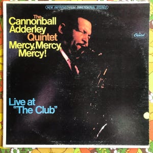 May include: A black and white vinyl record cover featuring a photo of a man playing a saxophone. The album is titled "The Cannonball Adderley Quintet Mercy, Mercy, Mercy! Live at "The Club"" and is a Capitol Records release.