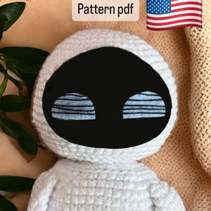 Puede incluir: Juguete amigurumi de crochet blanco que se asemeja a un robot con ojos negros y rayas azules. El juguete está sentado sobre una superficie marrón con una pequeña bandera estadounidense en el fondo. El texto "Pattern pdf" es visible en la parte superior de la imagen. El texto "Amigurumi_ak" es visible en la parte inferior de la imagen.