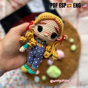 Puede incluir: Muñeca de ganchillo hecha a mano, con cabello rubio de hilo, ojos negros grandes y un mono azul con detalles de estrellas blancas. La muñeca tiene un tono de piel claro y lazos rojos. El texto de la imagen dice "PDF ESP ENG" y "@amigurumi_ak".