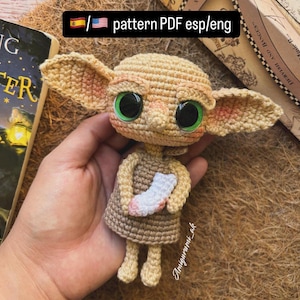 Puede incluir: Muñeca de ganchillo con grandes ojos verdes, orejas largas y un vestido pequeño, sosteniendo un calcetín blanco y rosa. La muñeca es de color beige y marrón, con el texto "pattern PDF esp/eng" en la esquina superior izquierda.