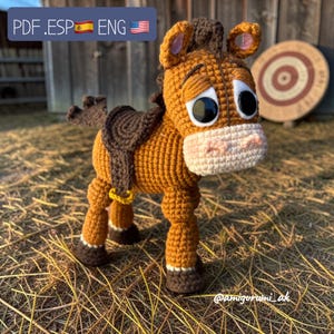 Puede incluir: Un juguete de caballo hecho a mano con ganchillo en tonos marrón, beige y blanco. El caballo tiene ojos grandes y expresivos, una silla de montar y un anillo amarillo. El texto "PDF .ESP ENG" está en la esquina superior izquierda. El juguete está sobre un fondo rústico.