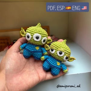 May include: Two crocheted green and blue alien toys with three eyes each. The aliens are wearing blue jumpsuits with yellow accents. The image includes the text "PDF. ESP ENG" and the Instagram handle "@amigurumi_ak".