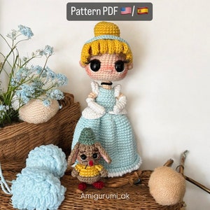Puede incluir: Una muñeca amigurumi de ganchillo de Cenicienta con un vestido azul con detalles blancos. La muñeca tiene el pelo rubio y ojos grandes. Un ratón amigurumi de ganchillo más pequeño con un sombrero verde y una camisa amarilla está sentado delante de la muñeca. El texto "Amigurumi_ak" está escrito debajo de las muñecas.