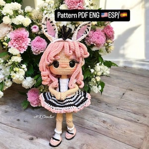 Puede incluir: Una muñeca de crochet con pelo rosa, un vestido blanco con rayas negras y blancas y orejas de conejo. La muñeca está de pie sobre una superficie de madera con flores rosas en el fondo. El texto "Pattern PDF ENG ESP/" es visible en la esquina superior derecha de la imagen.