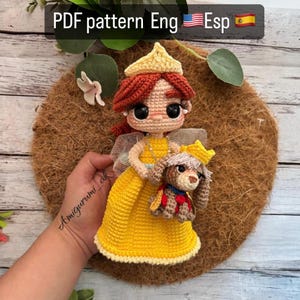 Puede incluir: Muñeca amigurumi de crochet con un vestido amarillo, cabello rojo y una corona. La muñeca sostiene un perro de crochet con un collar rojo y una corona. La muñeca está sobre un fondo marrón.