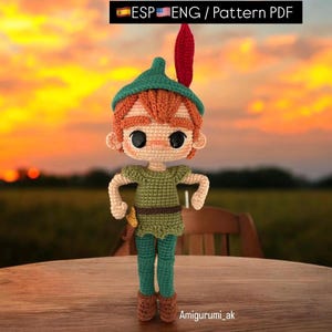 Puede incluir: Un juguete de amigurumi tejido a crochet de Peter Pan, un personaje ficticio de la novela Peter Pan y Wendy. El juguete está de pie sobre una mesa de madera con un fondo de puesta de sol. El juguete lleva una camisa verde, pantalones marrones y un sombrero verde con una pluma roja.