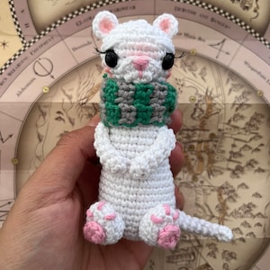 May include: White crocheted amigurumi ferret wearing a green and gray scarf. The ferret has pink ears, nose, and paw pads. The ferret is sitting on a hand, with a map in the background.