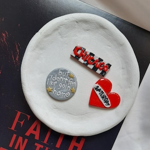 Può includere: Tre spille in argilla fatte a mano su un piatto bianco. Le spille sono un cuore rosso con la parola "Sabato" su di esso, un cerchio argentato con il testo "ma non ho voglia di tornare a casa", e una spilla rettangolare bianco e nero con la parola "Chicago" su di essa.