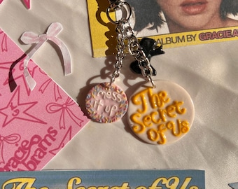 Puede incluir: Un llavero de metal plateado con un cierre de mosquetón y dos dijes circulares. Un dije es blanco con el texto "mess it up" y chispas de colores, mientras que el dije más grande es blanco con el texto amarillo "The Secret of Us".