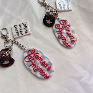 Può includere: Due portachiavi con chiusure color argento. Un portachiavi ha un ciondolo a forma di cappello da cowboy marrone e un'etichetta a scacchi blu e bianca con testo rosso in corsivo che recita "Kansas Thymeore". L'altro portachiavi ha un'etichetta con la scritta "Where's my Sally Tonight?"