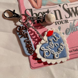 Op de afbeelding: Een zilverkleurige sleutelhanger met een blauwe hartvormige bedel met witte geschulpte randen, witte tekst Short n Sweet en een rood lippenicoon. Een tweede lichtblauwe rechthoekige bedel bevat de bruine tekst Espresso.