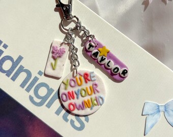 Puede incluir: Un llavero de metal plateado con tres dijes: uno rectangular blanco con una flor morada, uno circular blanco con el texto multicolor YOU'RE ON YOUR OWN KID y uno ovalado morado con el nombre TAYLOR y una estrella amarilla.