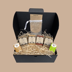 May include: A black gift box filled with natural soap bars and a soap net. The soaps are labelled with different scents, including "Classic Skin Care Soap", "Baby Skin Care Soap", "Talisman Skin Care Soap", "Vegan Skin Care Soap", "Milk & Honey Toothsoap", and "Vegan Toothsoap".
