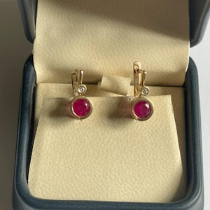 Puede incluir: Un par de pendientes de oro con cierre de palanca, cada uno con una piedra preciosa redonda de color rojo rosado vibrante engastada en bisel en la base y un pequeño diamante redondo transparente encima, presentados en un joyero azul oscuro.