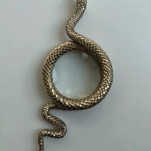 May include: A gold-colored magnifying glass with a snake design. The snake's body forms a circle around the glass, with the head and tail extending outwards. The snake has a textured, scale-like pattern.
