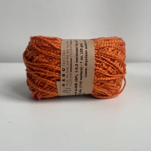 May include: A ball of orange yarn with a paper label wrapped around it. The label provides details about the yarn, including the brand, type, and care instructions. The yarn is approximately 28 grams and 116 metres.