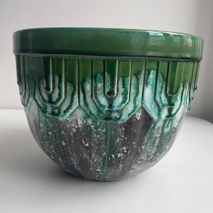 Op de afbeelding: Een ronde keramische plantenpot met een glanzende donkergroene rand en een textuur op het lichaam. Het bovenste gedeelte toont een herhalend geometrisch patroon, terwijl het onderste deel overgaat in een gevlekte donkergrijze en blauwgroene afwerking.