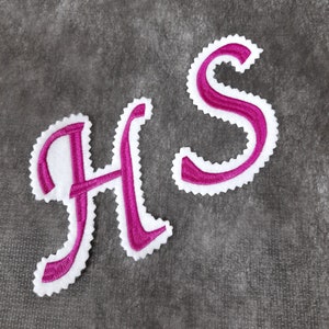 Könnte beinhalten: Zwei gestickte Buchstaben, "H" und "S", in einer rosa kursiven Schriftart mit weißen Umrandungen. Die Buchstaben befinden sich auf einem grauen strukturierten Hintergrund.