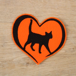 Könnte beinhalten: Eine schwarze Katzensilhouette in einem leuchtend orangefarbenen Filzherz. Das Herz ist schwarz umrandet.