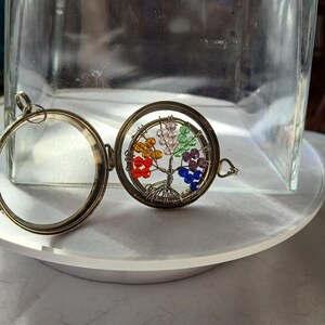 May include: Open, round, antique-style locket with a colorful tree of life design. The tree features red, orange, yellow, green, purple, and blue beads. The locket is made of metal and has a hinged opening.