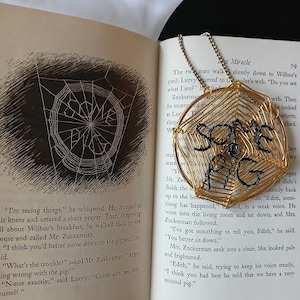 May include: A gold-coloured wire art bookmark with the words "SOME PIG" in black lettering, placed on the pages of a book. The book is open to a page with a black and white illustration of a spider web.