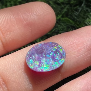 May include: An oval-shaped, faceted gemstone with a translucent purple base color and vibrant, multi-colored iridescent flecks of green, yellow, and blue light reflecting across its polished, smooth surface.