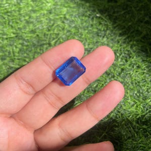 May include: A rectangular, faceted blue gemstone with a transparent appearance and sharp, clean edges. The stone displays a vibrant, uniform blue color throughout its structure, reflecting light across its polished, flat surfaces in a clear, geometric cut.
